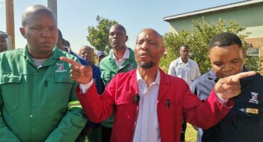 Men in work suits standing in front of a waste compactor truck at a depo.