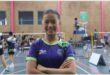 A dark-skinned girl with hair tied back in a purple and green shirt with her arms crossed with badminton court in the background.