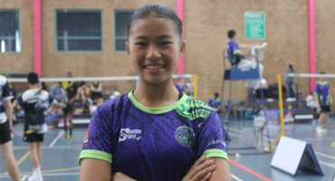 A dark-skinned girl with hair tied back in a purple and green shirt with her arms crossed with badminton court in the background.