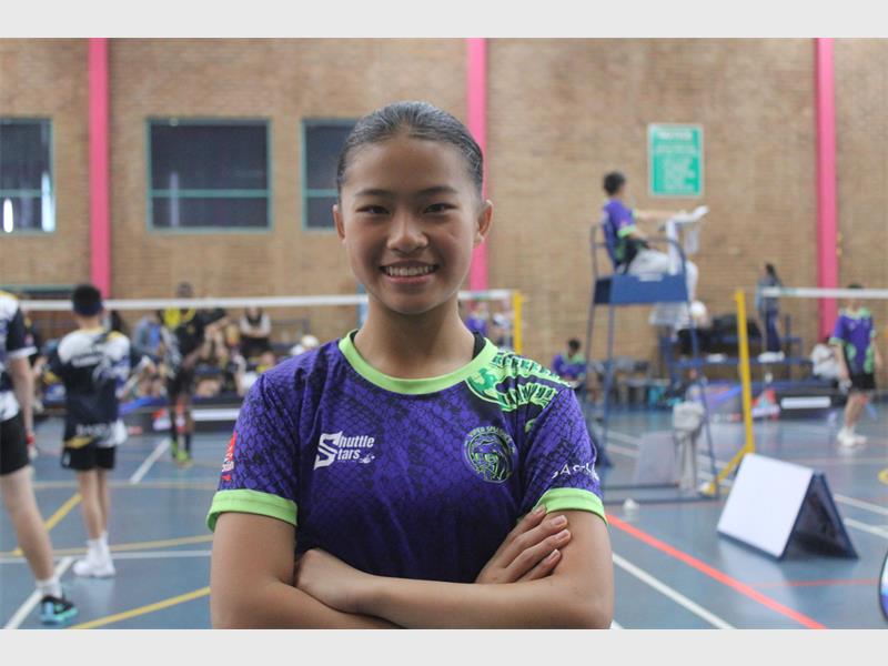 A dark-skinned girl with hair tied back in a purple and green shirt with her arms crossed with badminton court in the background.