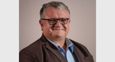 An elderly white male wearing glasses, a brown jacket and a blue shirt, smiling at the camera.