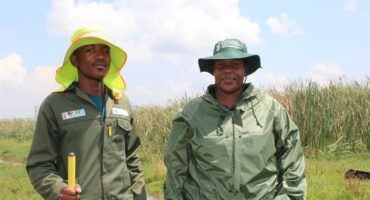 Two people a man and a woman in green work suits with the man holding a garden rake