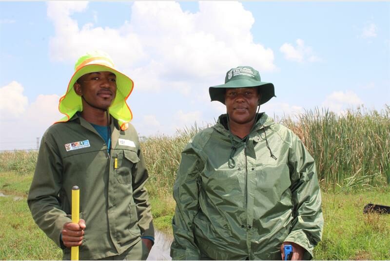Two people a man and a woman in green work suits with the man holding a garden rake