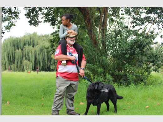 A man carrying a child on his shoulder and walking in a park with his on-leash dog