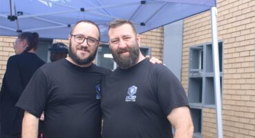 Two men in black t-shirts standing under a gazebo next to a building