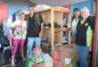 A group of four people in a clothing donation room, surrounded by shelves with folded clothes. Various canned and packaged food items on the floor add a community service vibe. Smiles and casual attire convey camaraderie and teamwork.