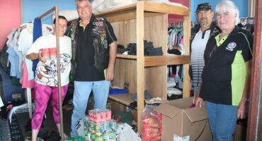 A group of four people in a clothing donation room, surrounded by shelves with folded clothes. Various canned and packaged food items on the floor add a community service vibe. Smiles and casual attire convey camaraderie and teamwork.