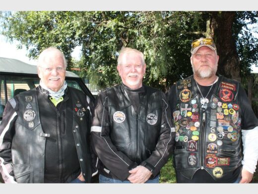 Three men in leather vests, adorned with various patches, stand outdoors smiling in front of a leafy tree. The atmosphere is friendly and relaxed.