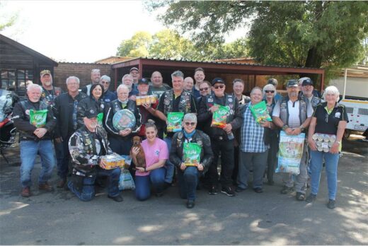A group of people wearing biker attire gather outdoors, smiling and holding pet food and supplies. The scene conveys camaraderie and charity support.