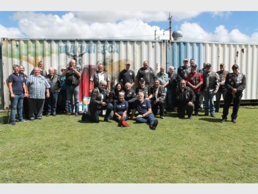 A group of diverse people poses happily on grass in front of a mural-painted container under a blue sky. The scene conveys camaraderie and community spirit.