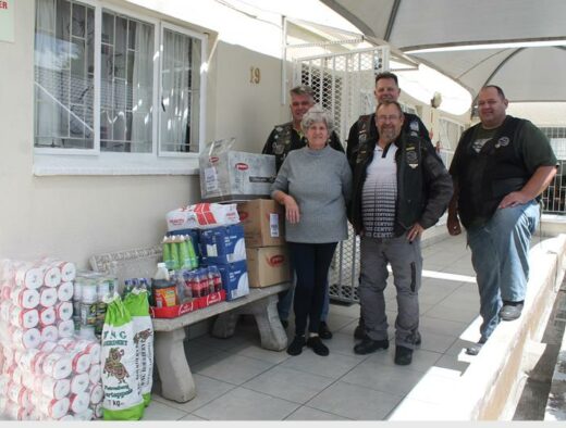 A group of five people stand smiling on a covered patio, surrounded by donations including toilet paper, canned foods.