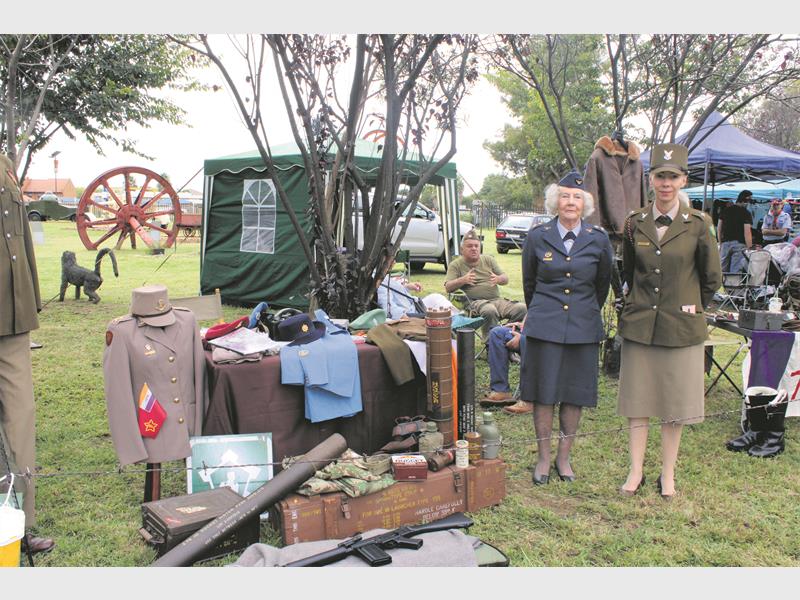 Two women in vintage military uniforms stand beside a display of historical military memorabilia, including uniforms, hats, and old equipment. The scene conveys a sense of nostalgia and historical appreciation.