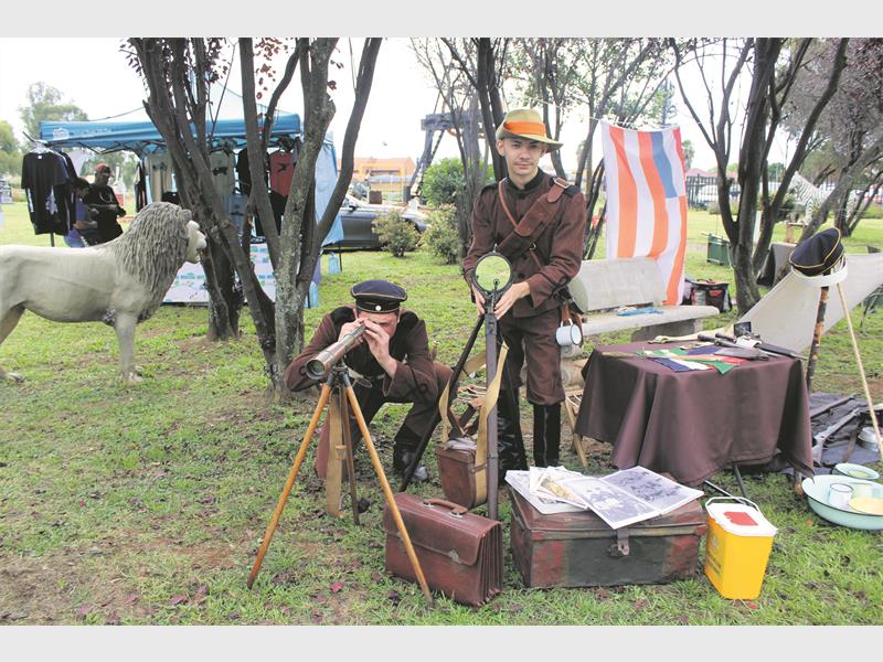 Two people in vintage explorer outfits are in a grassy area. One looks through a telescope, the other holds a magnifying glass. A lion statue and striped tent are in the background. The scene is colorful and playful.
