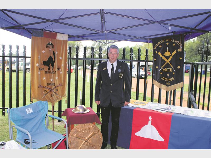 A man in a formal blazer stands under a blue canopy. He's surrounded by military memorabilia, banners, and a table with brochures, conveying respect and remembrance.
