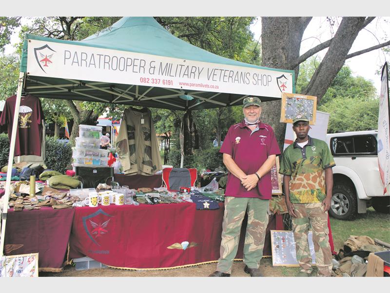 Two men in camouflage pants stand in front of a tent labeled "Paratrooper & Military Veterans Shop" with military-themed items on display.