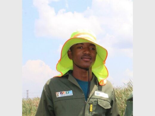 A black male wearing a yellow floppy had and a green overall top.