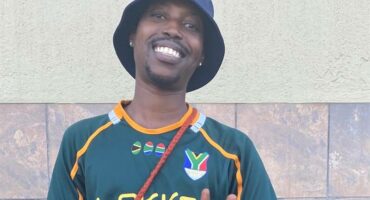 A black male smiling and wearing a Springbok shirt, a black bucket hat and giving a peace sign.
