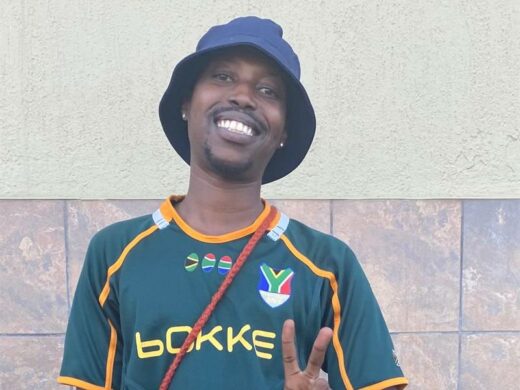 A black male smiling and wearing a Springbok shirt, a black bucket hat and giving a peace sign. 