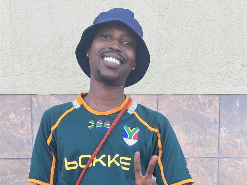 A black male smiling and wearing a Springbok shirt, a black bucket hat and giving a peace sign.
