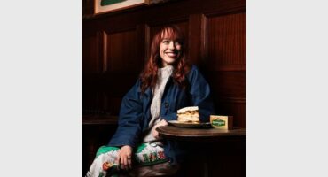 A red-haired woman dressed in light clothing and a denim jackets sitting at a small wooden table in a pub with a crisp sandwich and block of Kerrygold butter next to her.