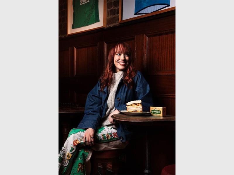 A red-haired woman dressed in light clothing and a denim jackets sitting at a small wooden table in a pub with a crisp sandwich and block of Kerrygold butter next to her.