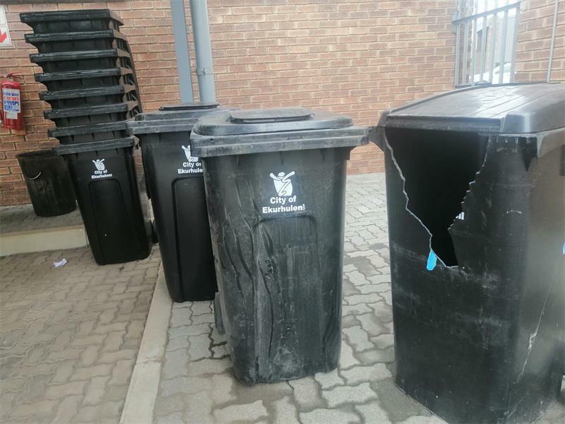 An image of black City of Ekurhuleni wheelie bins, some stacked others standing single and one broken.