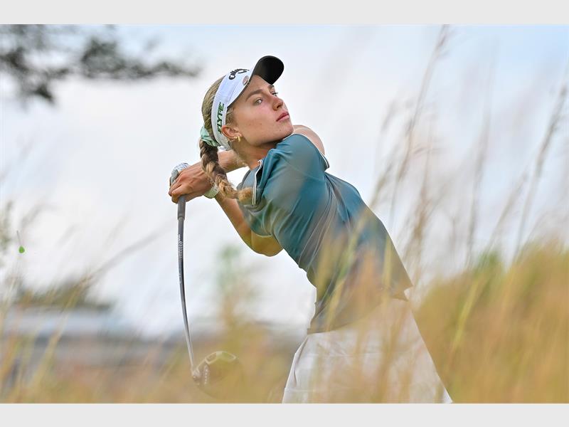 A female golfer completing a swing, wearing a blue shirt, white skirt and white cap.