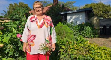A woman in red pants and floral top standing next to flowers and greenery