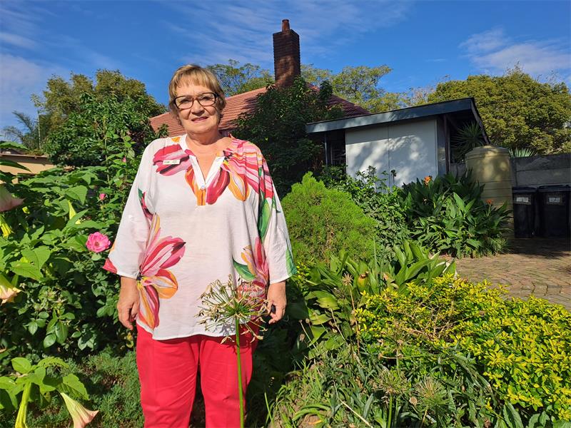 A woman in red pants and floral top standing next to flowers and greenery