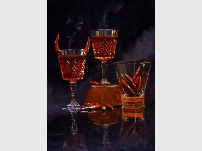 A photograph taken against a dark background of three wine glasses with red wine in them and chillies in the one glass as well as among the glasses on the table.