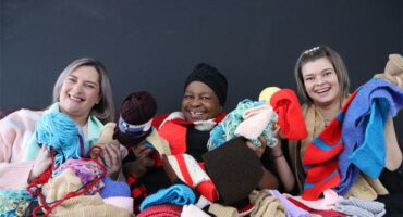 Three women - two white and one black - wrapped in woolen scarves and blankets and holding balls of wool.