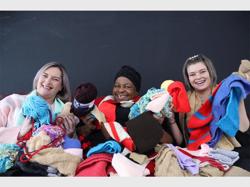 Three women - two white and one black - wrapped in woolen scarves and blankets and holding balls of wool.