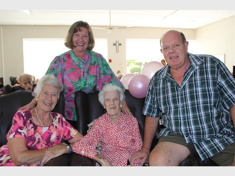 Two women and a man, all white, seated around an armchair in which a white woman celebrating her 101st birthday is seated wearing a red and white dress.