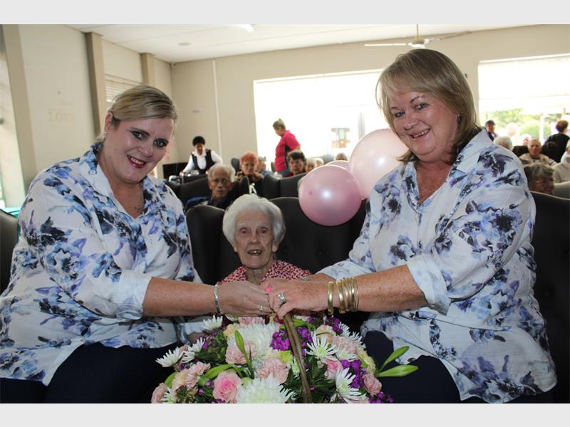 Two white sisters from a nursing home sitting on the armrests of a chair in which a lady celebrating her 101st birthday is seated wearing a red and white dress.