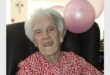 A white woman smiling as she celebrates her 101st birthday with two pink balloons tied to the chair she is sitting in.