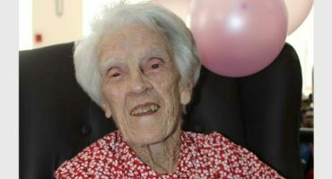 A white woman smiling as she celebrates her 101st birthday with two pink balloons tied to the chair she is sitting in.