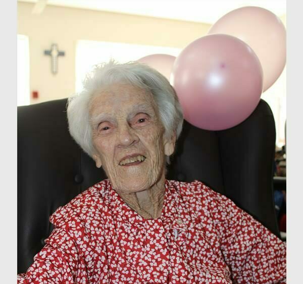A white woman smiling as she celebrates her 101st birthday with two pink balloons tied to the chair she is sitting in.