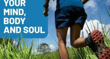 An image taken from behind of a man running in long green grass wearing shorts, a T-shirt and running shoes with the wording Excercise your mind, body and soul.