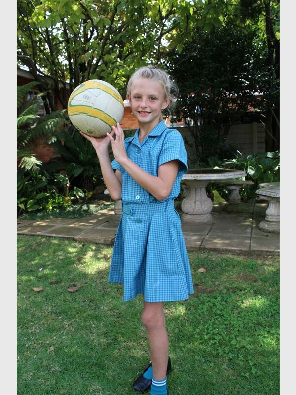 A young blonde girl in a blue school uniform holding a netball ball. 