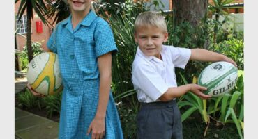 A blonde girl in a blue school uniform holding a netball ball and a smaller blond boy in a white school shirt and grey shorts holding a rugby ball.
