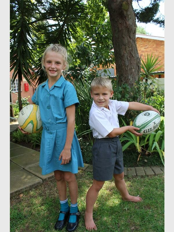 A blonde girl in a blue school uniform holding a netball ball and a smaller blond boy in a white school shirt and grey shorts holding a rugby ball.