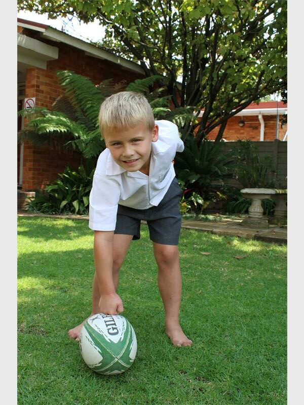 A young blond boy in grey school shorts and a white school shirt standing on a lawn and reaching down with one hand to a rugby ball at his feet. 