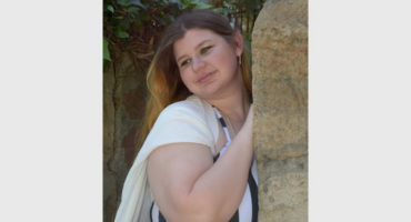 A young girl with long dark hair smiling and leaniing up against a tree looking away from the camera wearing a black and white top and black skirt.