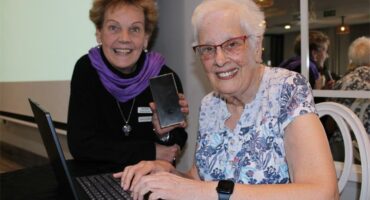 Two women, one dressed in black with a purple scarf and the other in short-sleeve dress with grey hair and glasses working on a laptop.