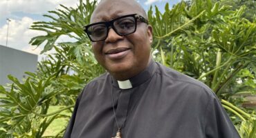 A black male wearing a clergy black shirt with a cross around his neck and black rimmed glasses.