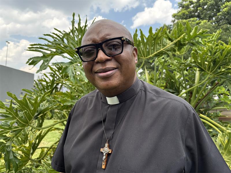 A black male wearing a clergy black shirt with a cross around his neck and black rimmed glasses.