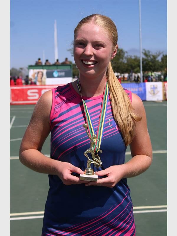 A netball player photographed on a court with long blonde hair in a blue and pink kit holding a medal that is around her neck.