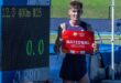 A young white athlete in purple kit holding a red piece of paper with the word National in white and standing next to a time board on an athletics track.