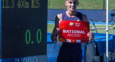 A young white athlete in purple kit holding a red piece of paper with the word National in white and standing next to a time board on an athletics track.