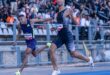 Two male athletes in action sprinting across the finish line with the crowd in a stadium in the background.